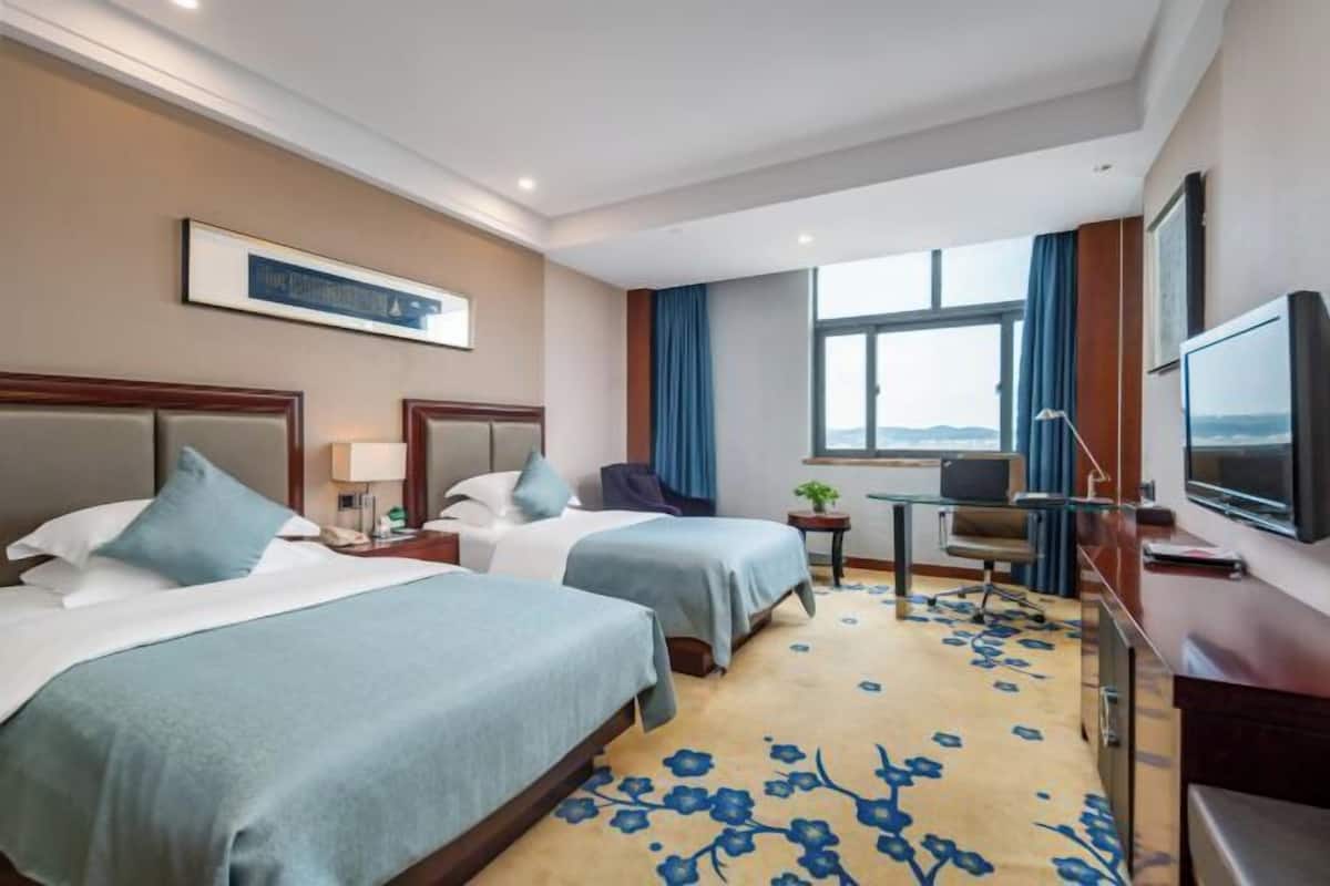A hotel room with two beds, a desk, a chair, a TV, and a window with a view.