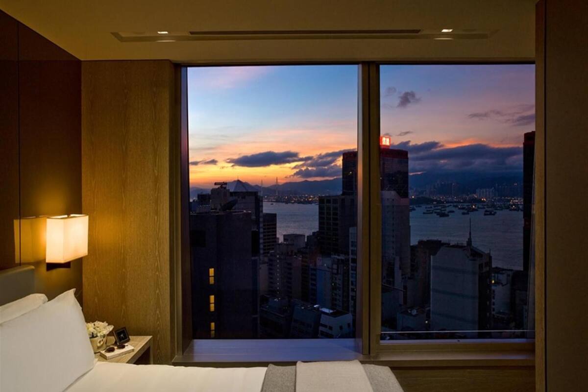 A hotel room with a large window offering a cityscape view at sunset.
