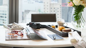 A desk with a camera, a book, a pair of glasses, a notebook, a pen, a cup, a plate with food, a vase with flowers, and a window with a city view. - One96 (Hong Kong)