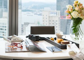 Un escritorio con una cámara, un libro, un par de gafas, una libreta, un bolígrafo, una taza, un plato con comida, un florero con flores y una ventana que ofrece vistas de la ciudad.