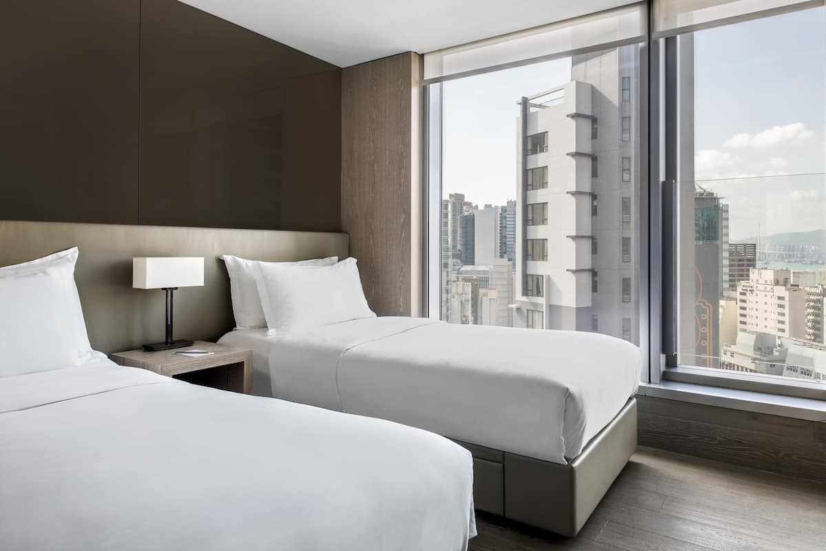 A hotel room with two beds, a nightstand, a lamp, and a large window offering a city view.