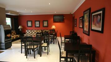Un comedor con pared roja, varias mesas y sillas, un televisor montado en la pared y cuadros enmarcados.