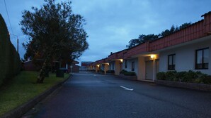 A street view of a motel with a row of single-story buildings, each with a garage door and a small front yard. - Motel Aeropuerto (Mos)