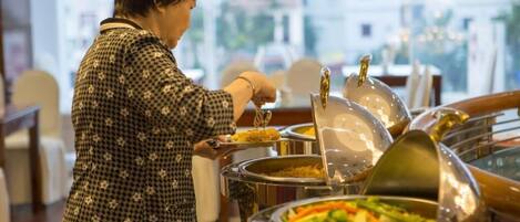 A buffet setup with various food items in metal containers, a person serving food, and a view of buildings through large windows.