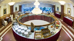A buffet setup with a central serving area, surrounded by dining tables and chairs, in a well-lit room with a chandelier. - Adina Hotel (Da Nang)