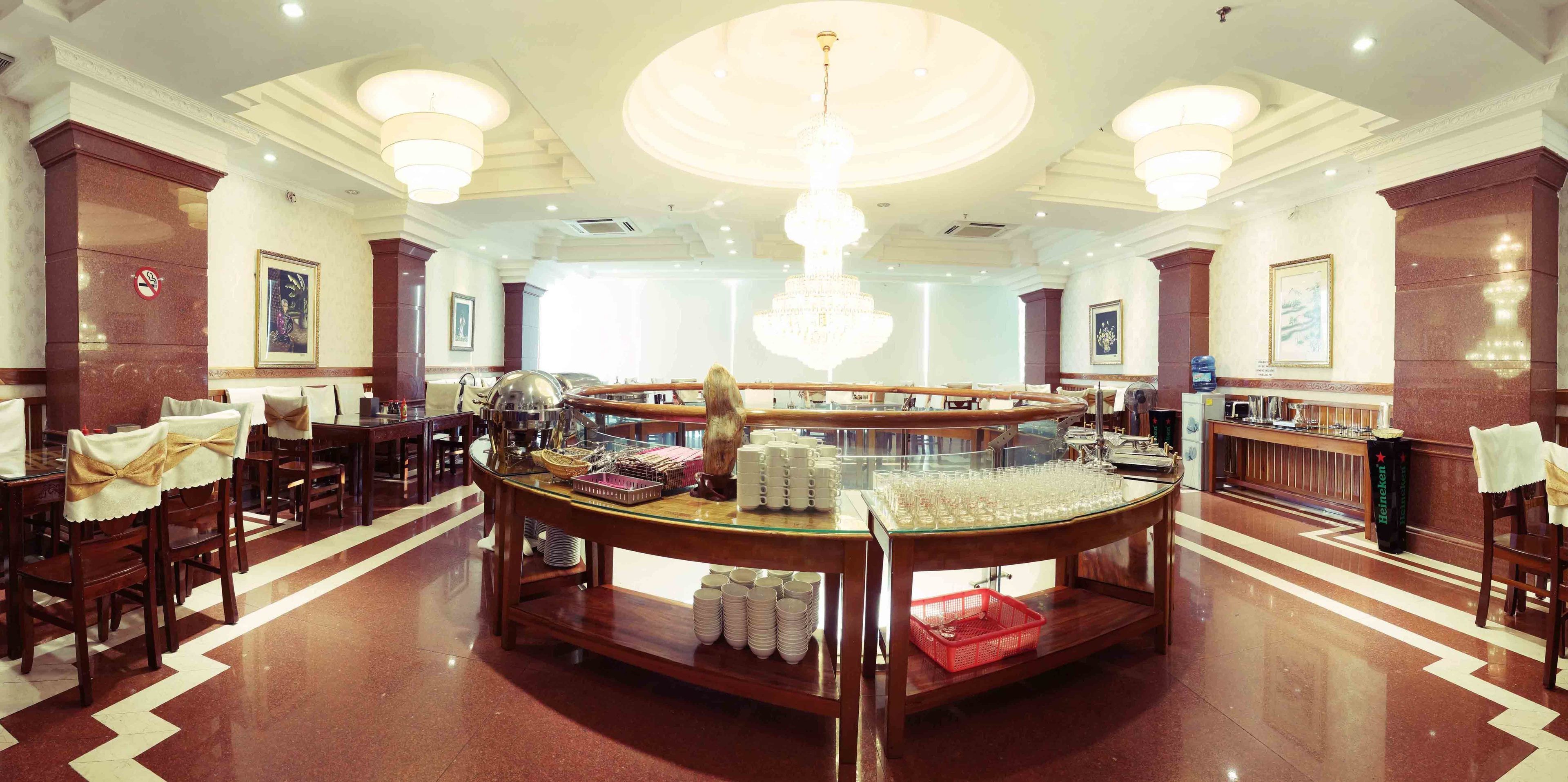 A hotel lobby with a central buffet area, wooden chairs with bow ties, and a large chandelier.