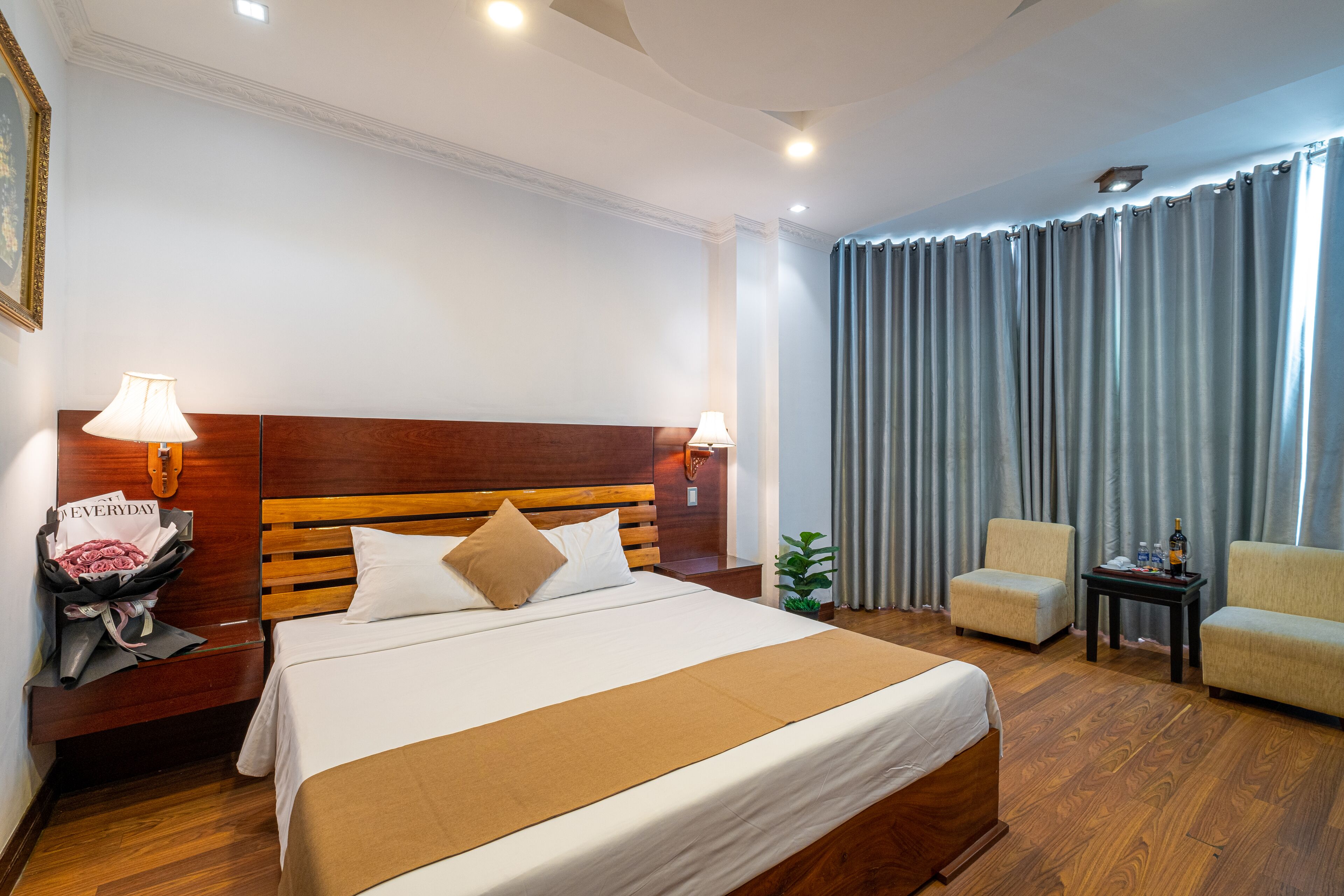 Ein Hotelzimmer mit Bett, Holz-Kopfteil, zwei Nachttischlampen, einem kleinen Tisch mit einer Flasche und einem Stuhl.