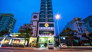 A hotel named Adina with a prominent sign, a modern building facade, and a street view with parked cars and a cafe. - Adina Hotel (Da Nang)