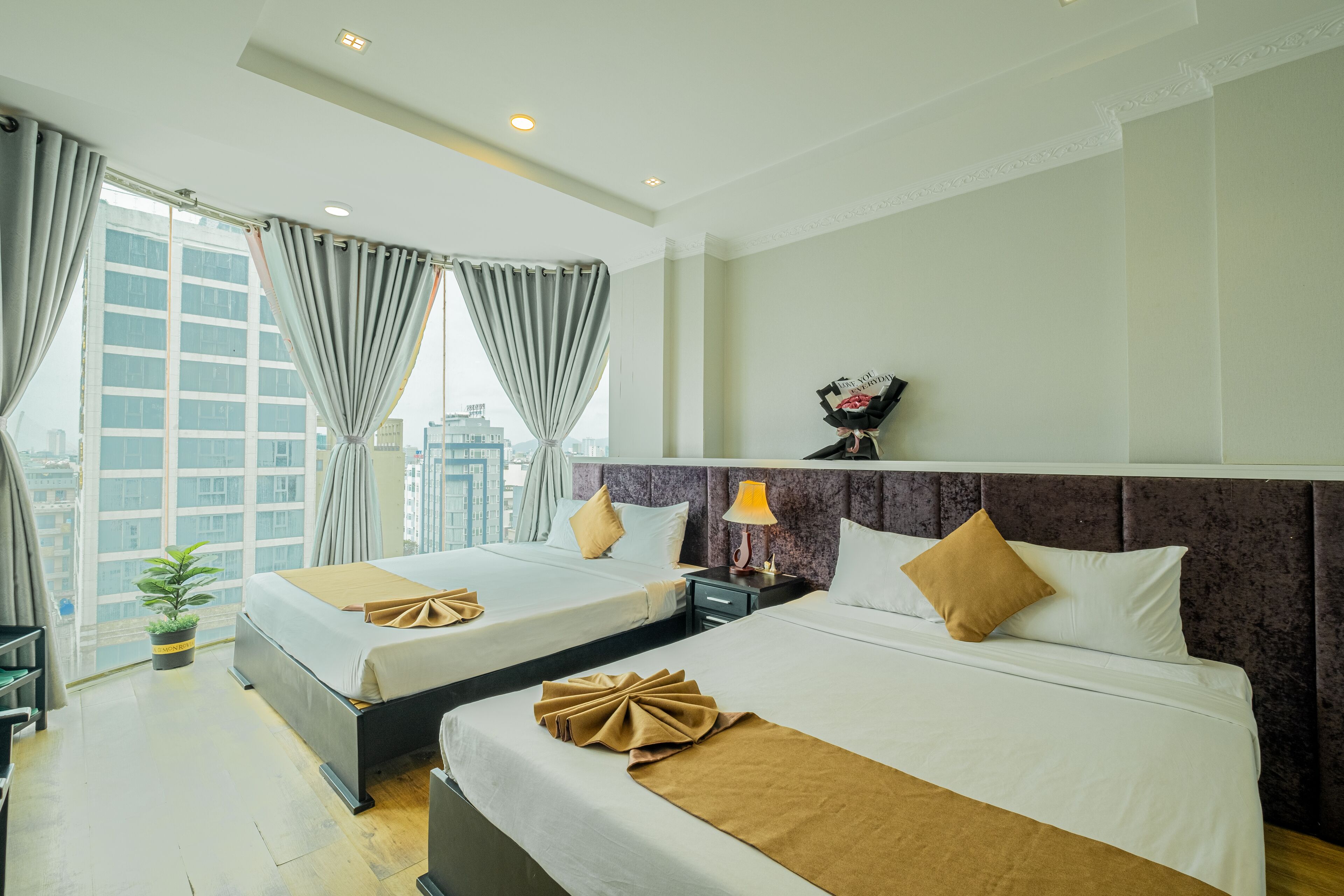 A hotel room with two beds, a large window with curtains, a small table with a lamp, and a potted plant.