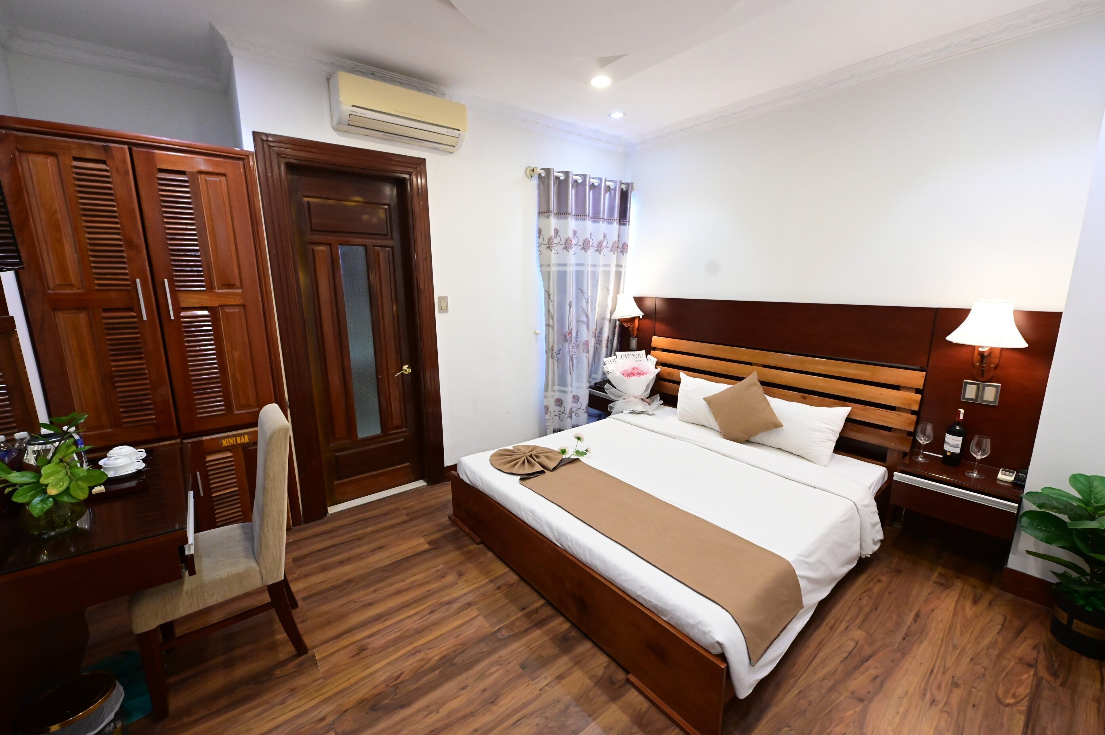 A hotel room with a wooden bed, a bedside table with a lamp, a nightstand, a desk with a chair, and a wardrobe.