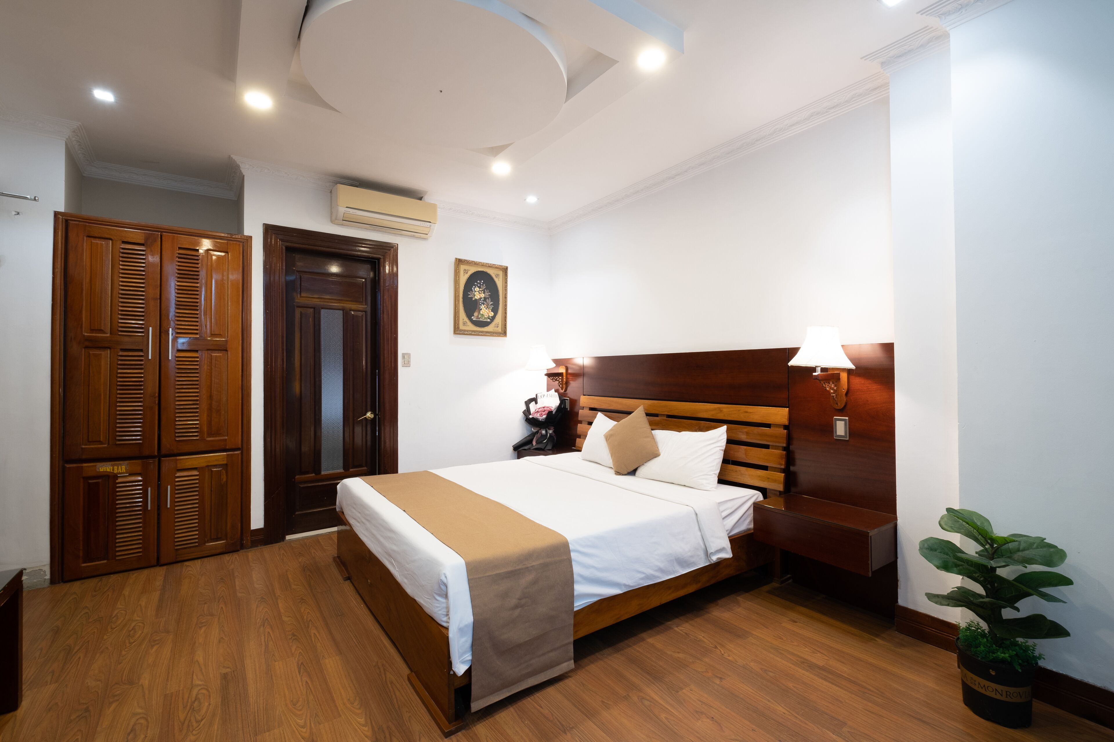 Une chambre d’hôtel avec un lit en bois, un banc, une plante en pot, une armoire et une lampe fixée au mur.