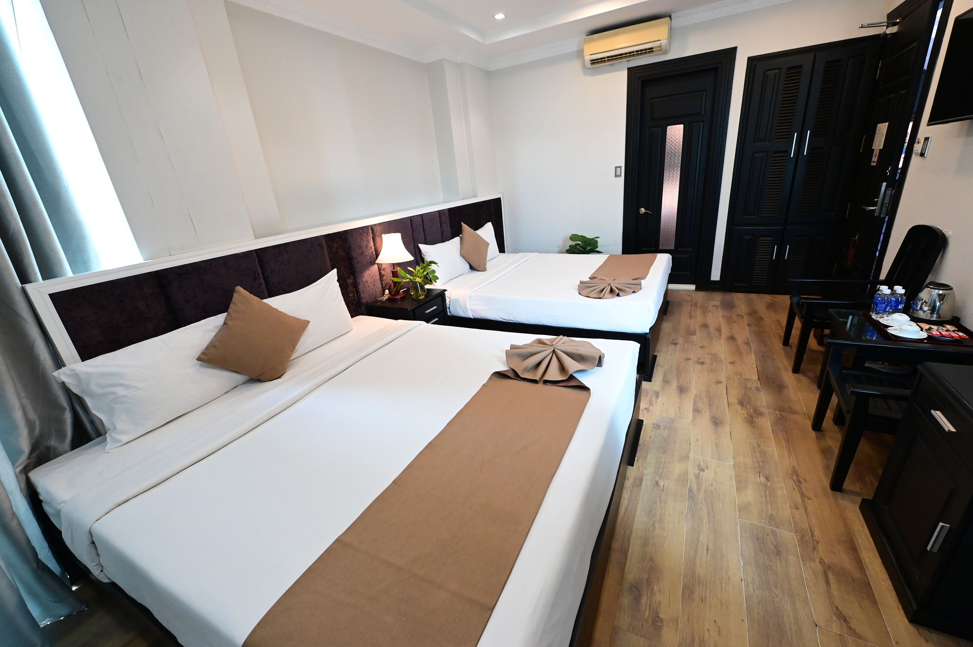 A hotel room with two beds, a wooden floor, a desk with a chair, and a wardrobe.