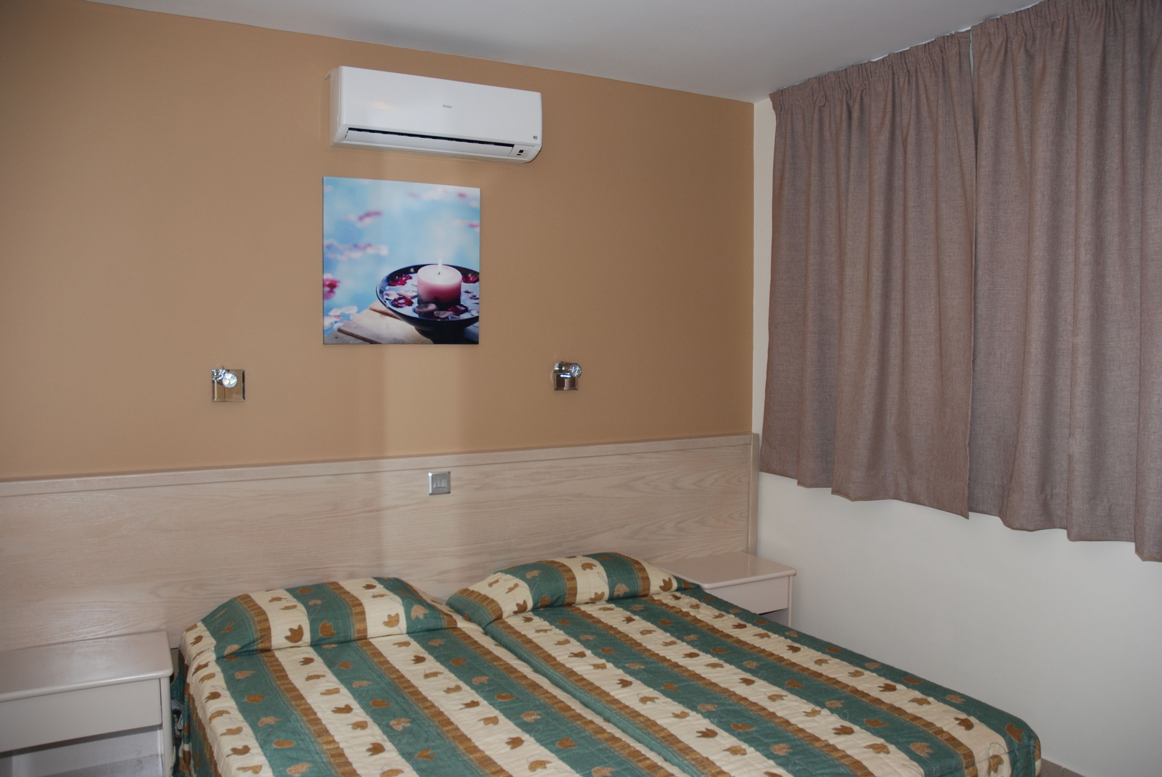 Una habitación de hotel con cama, mesita de noche, unidad de aire acondicionado y un cuadro en la pared.