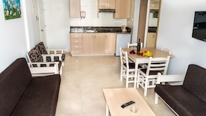 A compact living space with a sofa, a dining table with chairs, a kitchen area with cabinets and a dishwasher, and a TV mounted on the wall. - Christabelle Hotel Apartments (Ayia Napa)