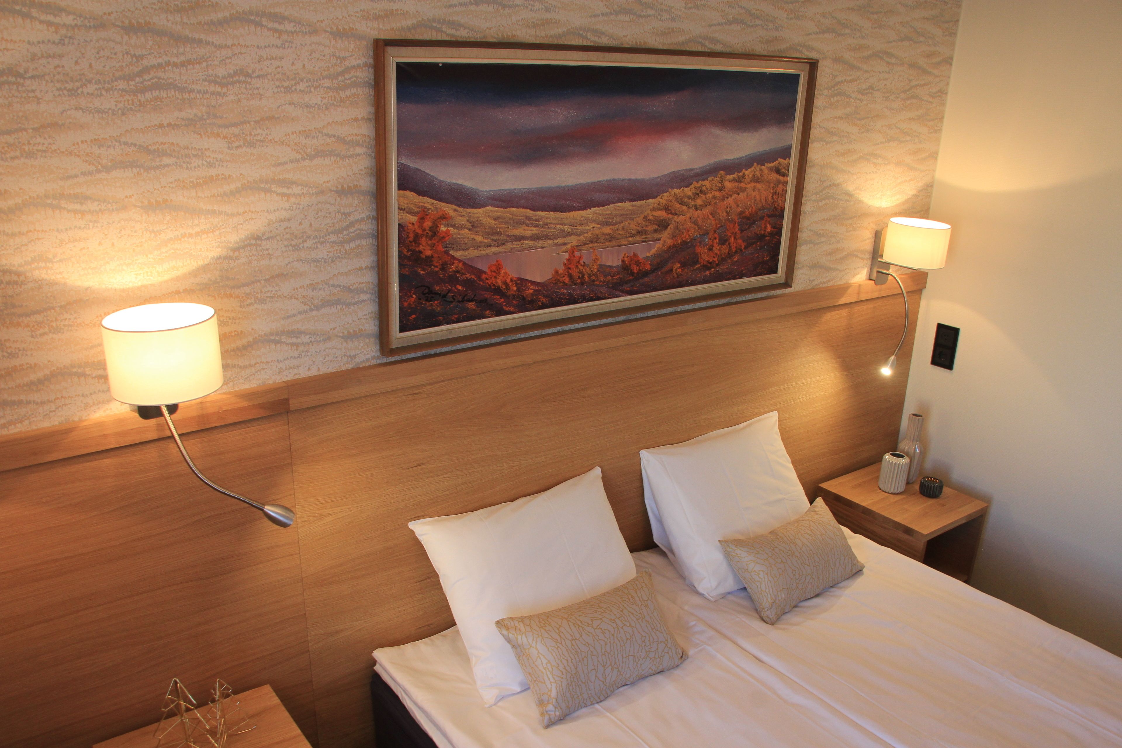 A neatly made bed with white linens and two pillows, flanked by two bedside lamps on wooden nightstands. A framed landscape painting hangs above the bed.