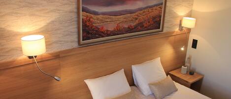 A neatly made bed with white linens and two pillows, flanked by two bedside lamps on wooden nightstands. A framed landscape painting hangs above the bed.