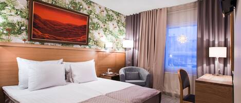 A hotel room with a bed, a desk, a chair, a nightstand, a lamp, a window with curtains, a wall with floral wallpaper, and a painting above the bed.