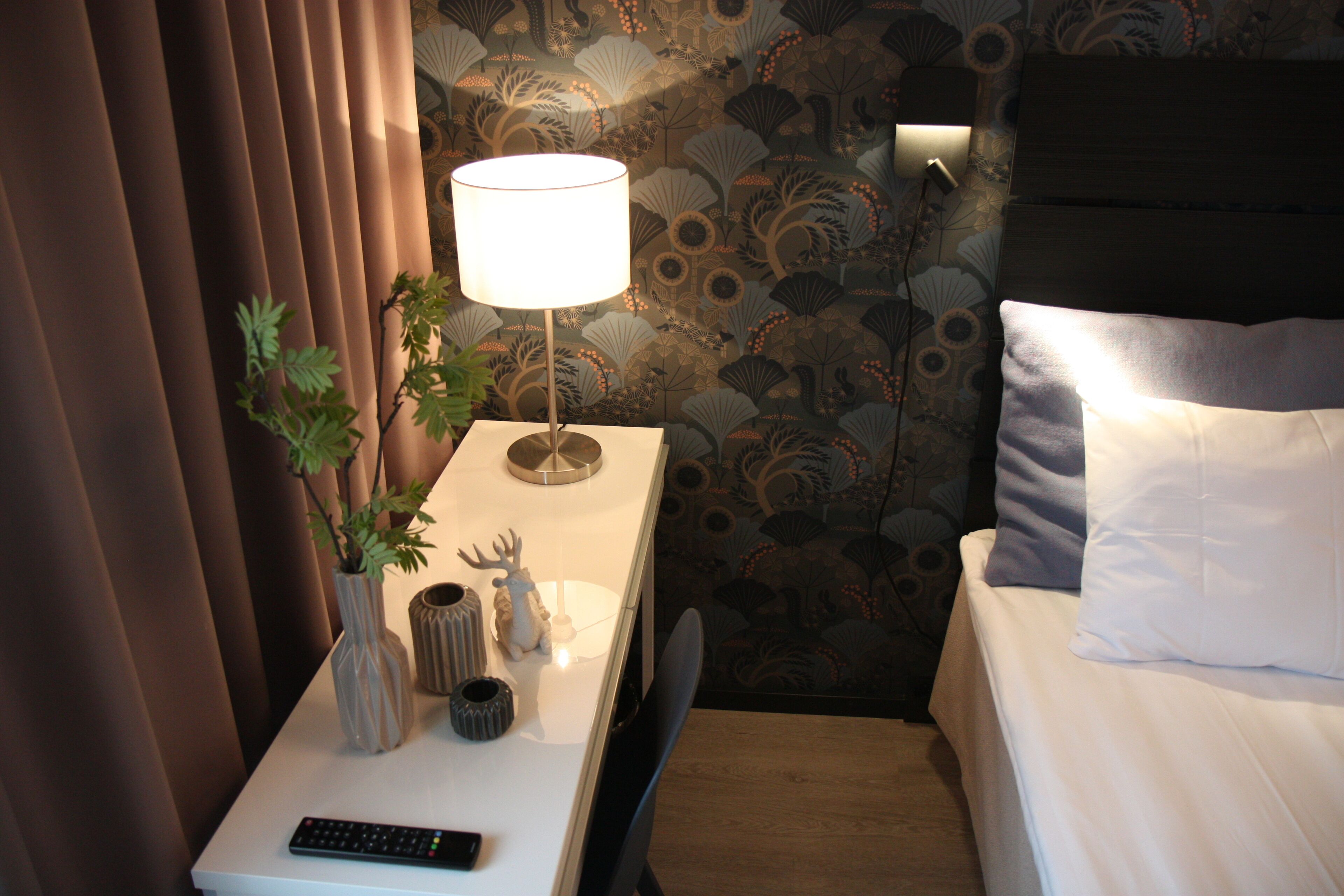 A neatly made bed with white linens, a bedside lamp, a remote control, and a small plant on a bedside table.