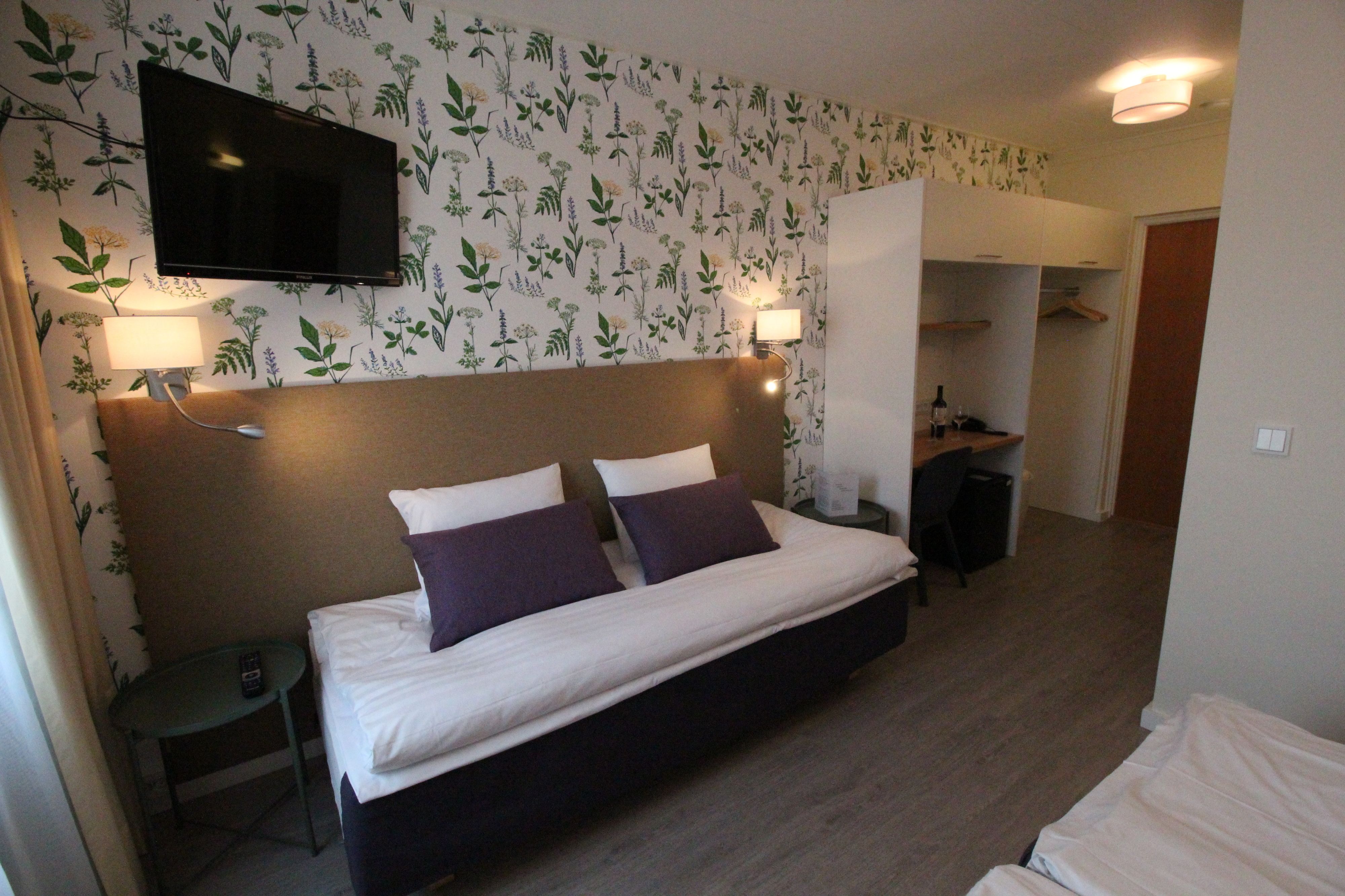 A hotel room with a bed, two bedside lamps, a TV mounted on the wall, and a small table with a bottle and a remote control.