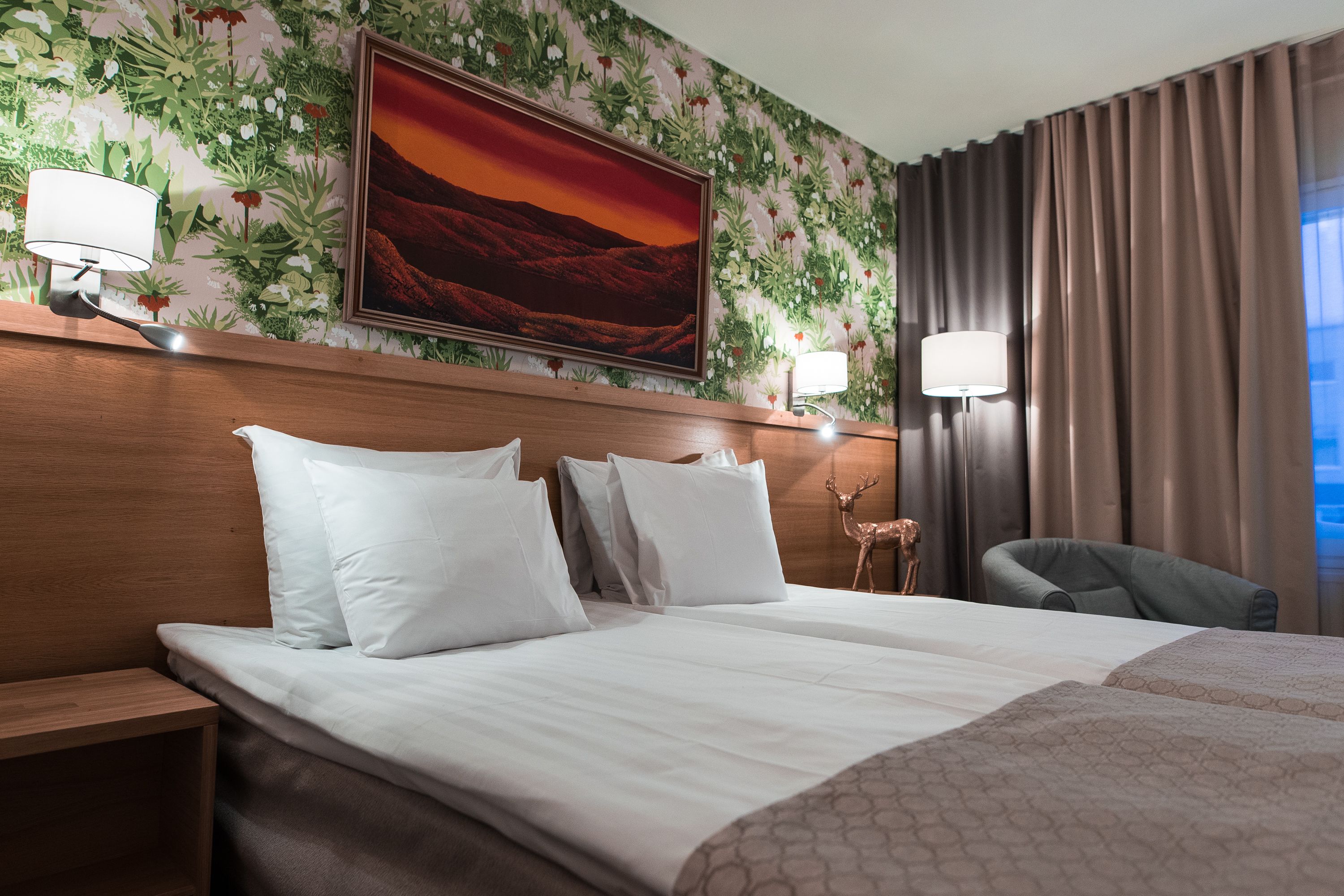 A hotel room with a large bed, two bedside tables, a nightstand, a chair, a wall-mounted lamp, and a painting of a sunset.