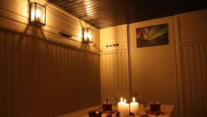 Un tavolo in legno con candele e bicchieri di una bevanda, posto di fronte a una parete con pannelli verticali e un quadro.