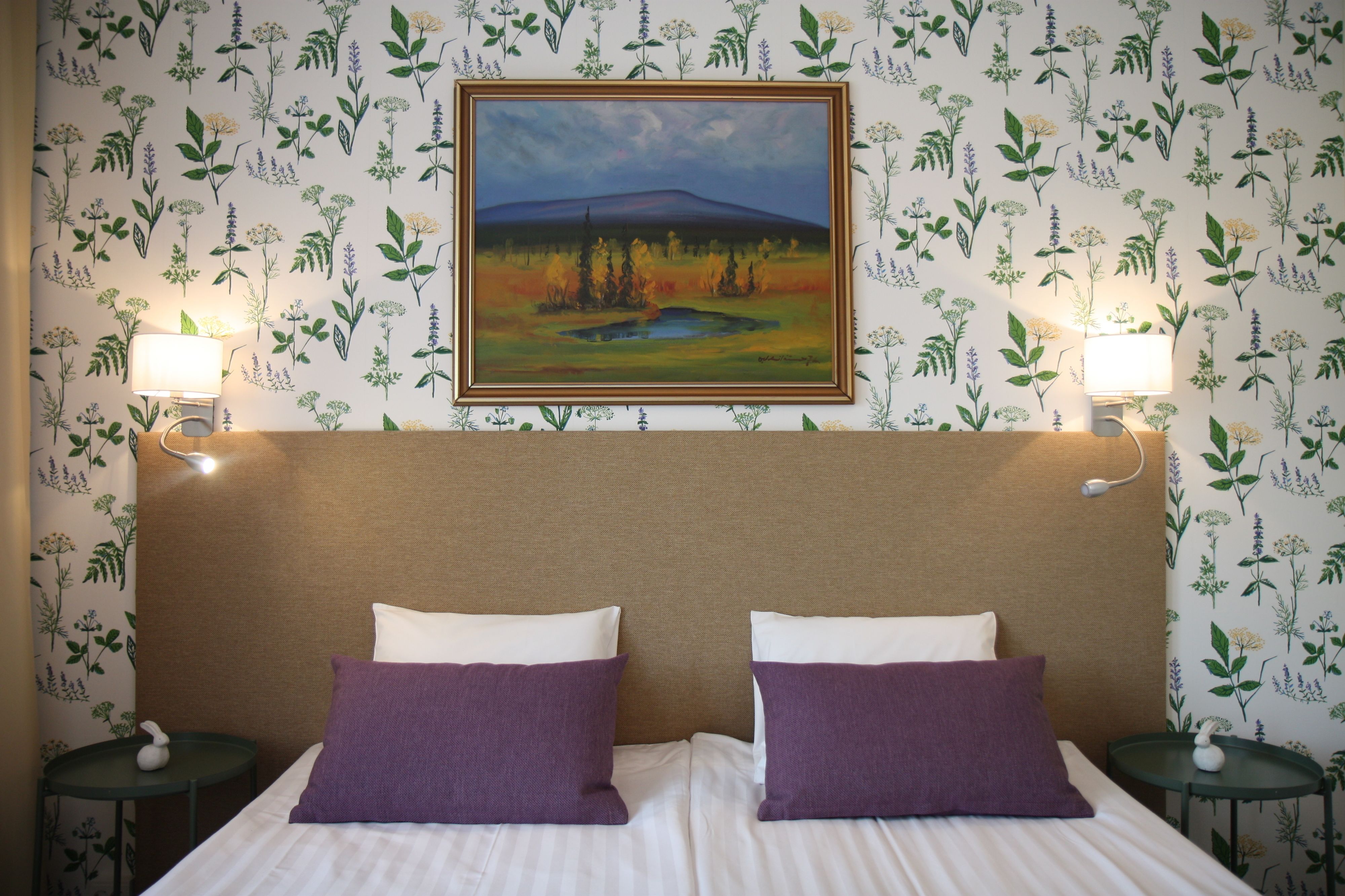 Una habitación de hotel con una cama, dos almohadas moradas, un cuadro de un paisaje y papel tapiz con motivos florales.