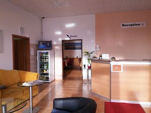 A reception area with a reception desk, a clock, a TV screen, a vending machine, and a seating area with a sofa and chairs. - Hotel New Fair Munich Messe (Munich)