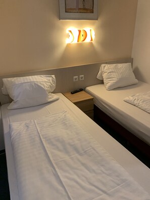 Habitación de hotel con dos camas individuales, ropa de cama blanca, una mesita de noche con control remoto y una lámpara fijada a la pared.