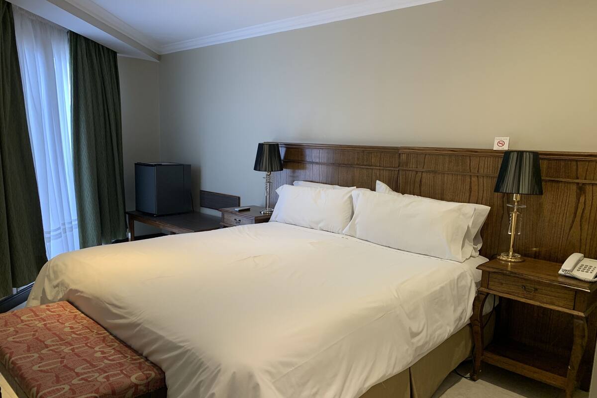 Une chambre d’hôtel avec un grand lit, une table de chevet avec une lampe, un téléphone, une petite table avec un téléviseur et un banc.