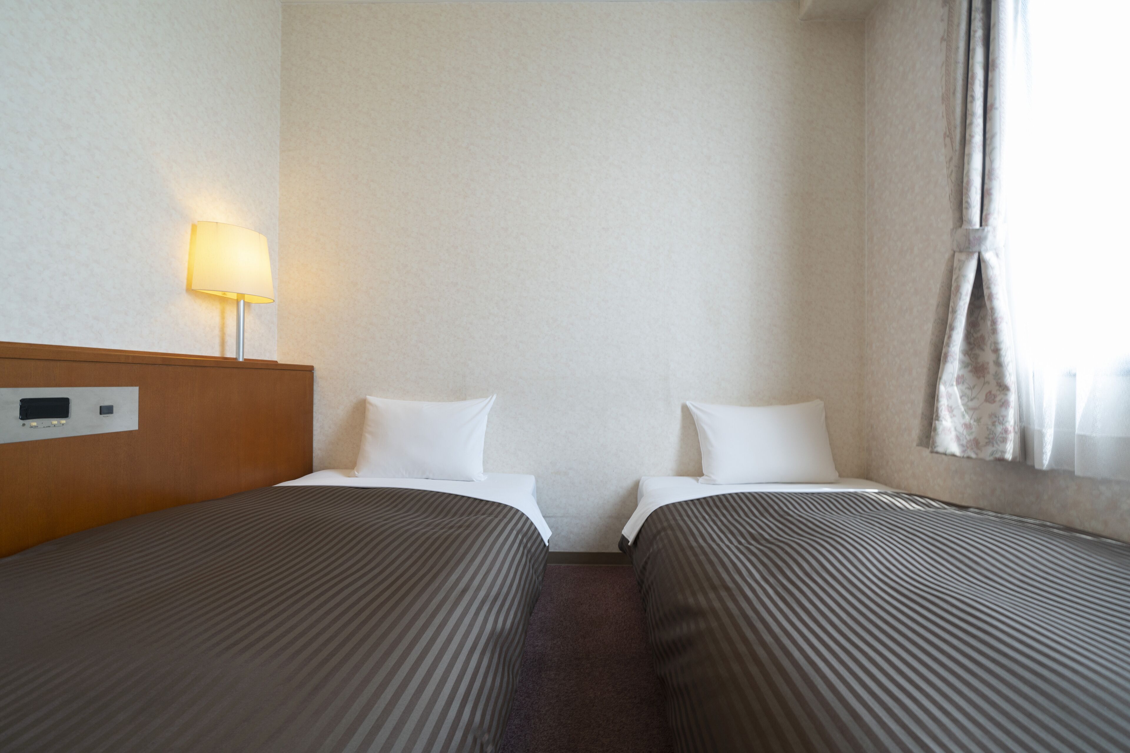 Une chambre d’hôtel avec deux lits, une tête de lit dotée d’un panneau de commande, une lampe et une fenêtre avec des rideaux.
