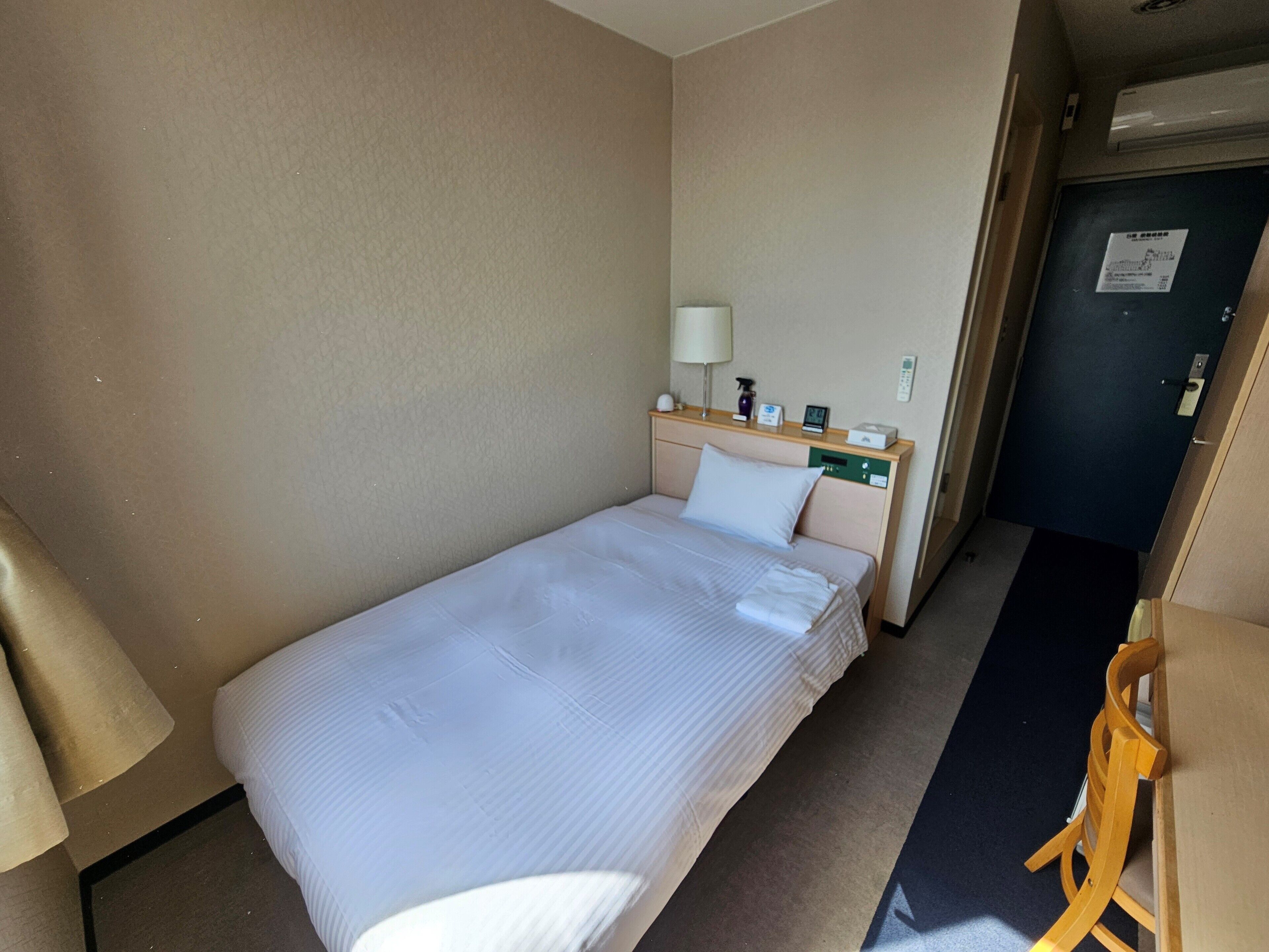 A single bed with a white comforter and pillows, a bedside table with a lamp, a small clock, and a control panel.