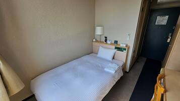 A single bed with a white comforter and pillows, a bedside table with a lamp, a small clock, and a control panel.