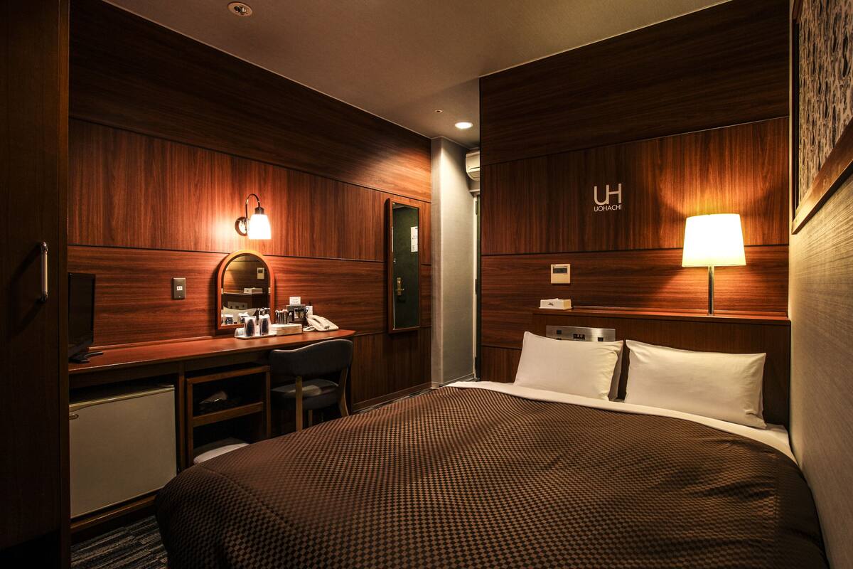 Une chambre d’hôtel avec un mur lambrissé en bois, un bureau avec une chaise, un lit recouvert d’une couvre-lit à motifs, une table de chevet et une lampe fixée au mur.