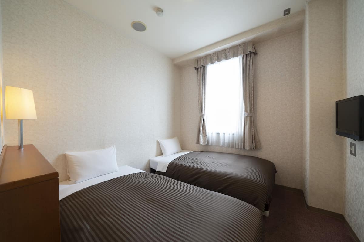 Une chambre d’hôtel avec deux lits, un bureau, une lampe, une télévision et une fenêtre avec des rideaux.
