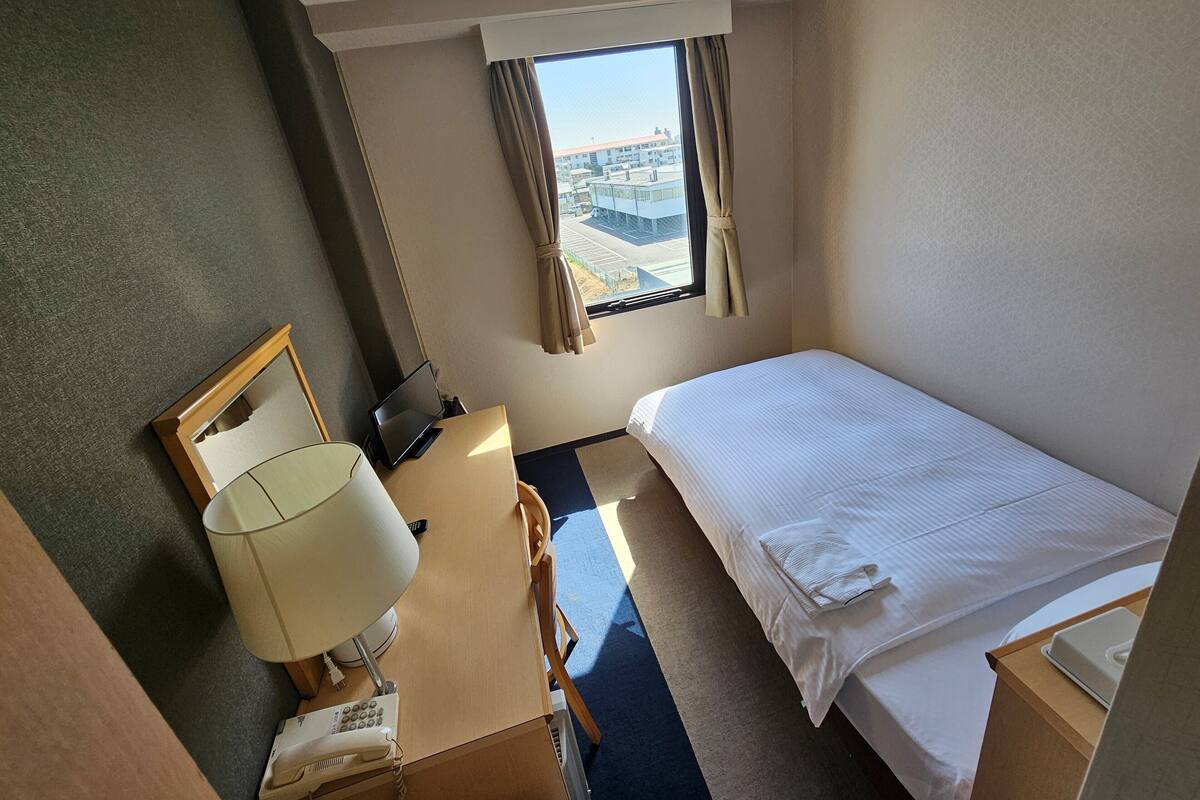Une chambre d’hôtel avec un lit, un bureau avec une lampe, un téléphone et une fenêtre avec des rideaux.