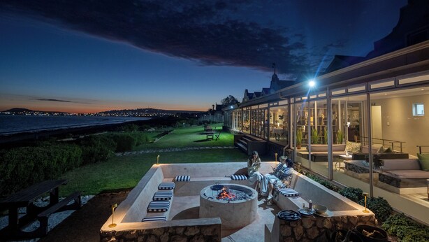 A well-lit outdoor patio with a fire pit, seating, and a view of the ocean at dusk. - Blue Bay Lodge (Saldanha)