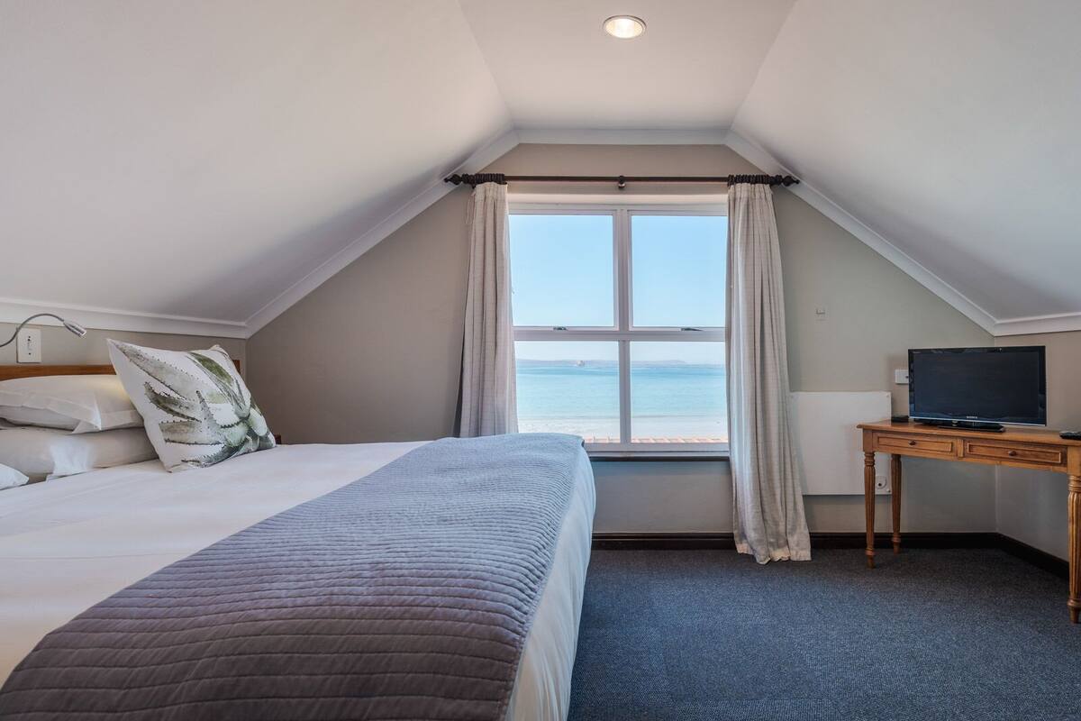 A bedroom with a bed, a desk with a television, and a window with a view of the sea.