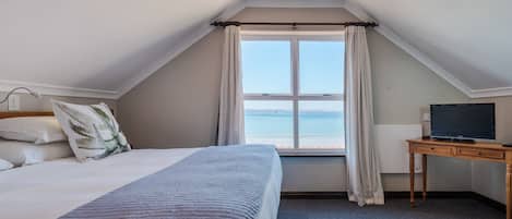 A bedroom with a bed, a desk with a television, and a window with a view of the sea.