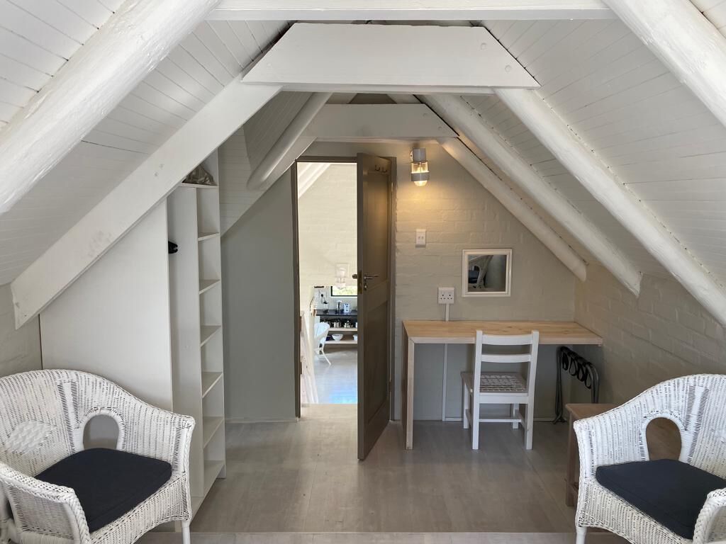 A cozy attic bedroom with a sloped ceiling, a small desk, two wicker chairs, and a view into the dining area.