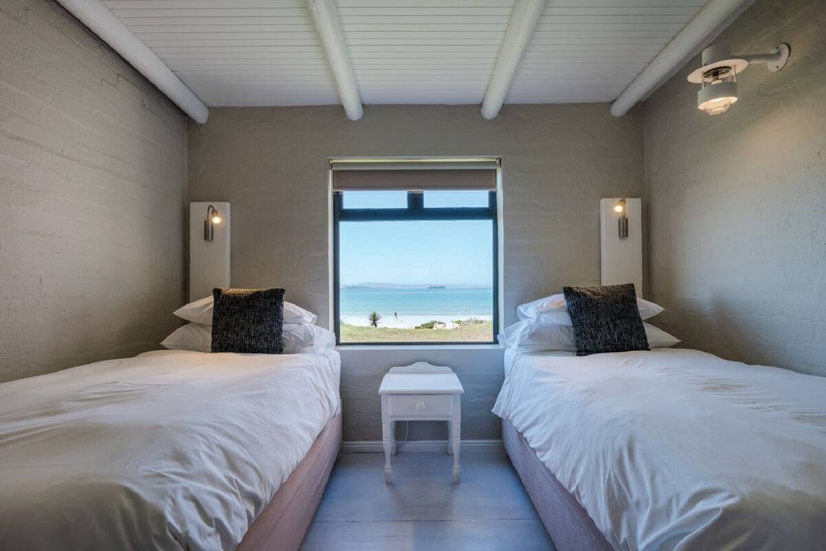 A bedroom with two beds, a small table, and a window with a view of the beach.