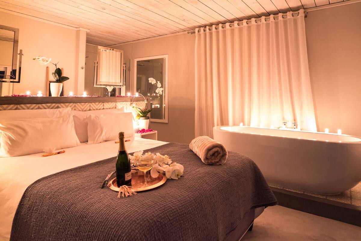 A hotel room with a large bed, a bathtub, and a bedside table with a bottle of champagne and flowers.