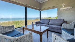 A modern room with a large glass window overlooking a beach, a grey sofa, wicker chairs, and a wooden table. - Blue Bay Lodge (Saldanha)