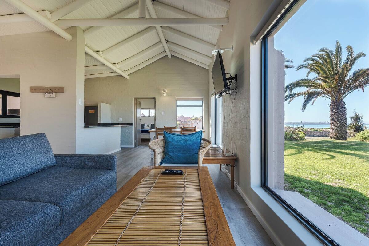 A spacious living area with a sofa, a wooden coffee table, and a view of a lush garden and ocean through large windows.