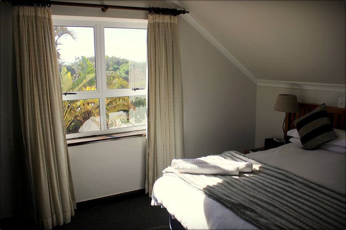 A bedroom with a large window, a bed, a nightstand, and a lamp.