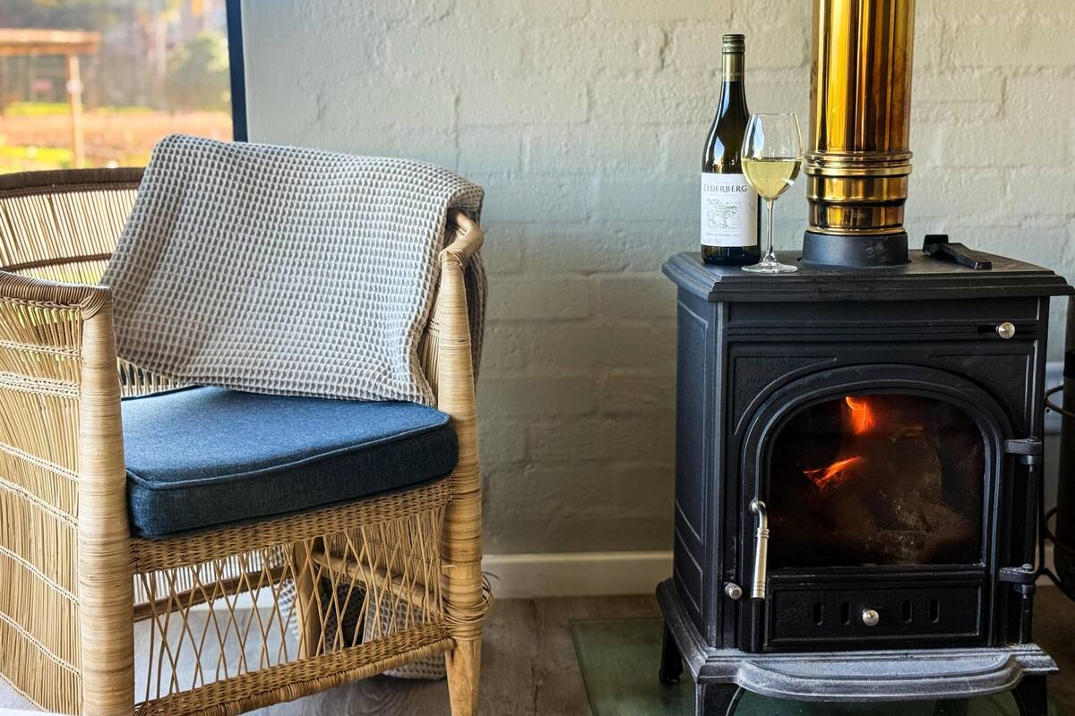 A wicker chair with a blue cushion, a bottle of wine, and a glass on a black wood-burning stove.