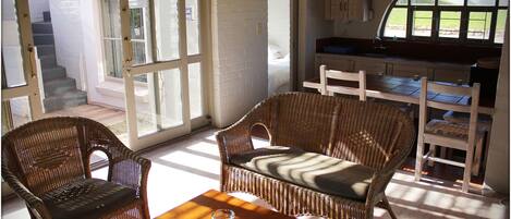 Une pièce meublée de meubles en osier, avec une table sur laquelle sont posées des télécommandes, et d’où l’on peut voir un escalier et une fenêtre.