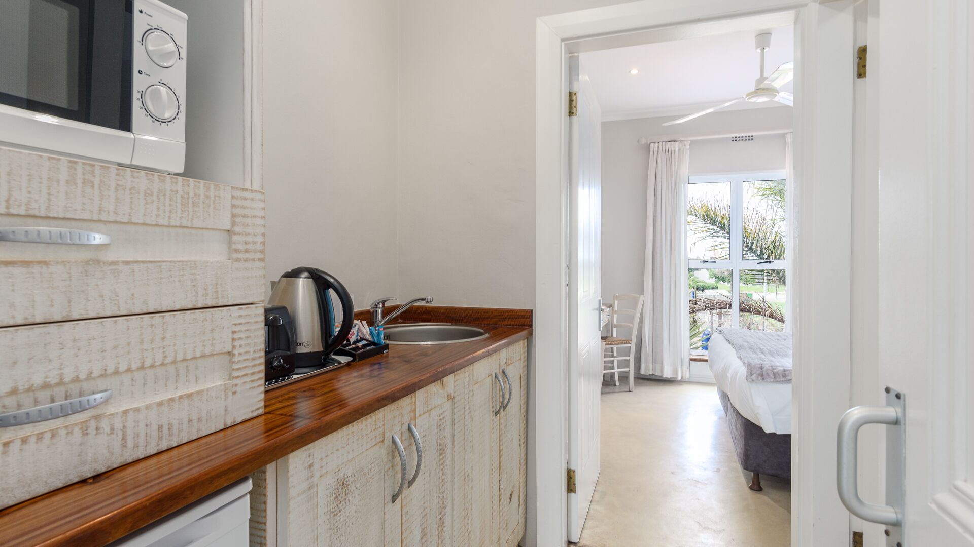 A compact kitchen with a microwave, kettle, and sink. A wooden counter and cabinets are visible, leading to a bedroom with a bed and a view of a garden through a window.