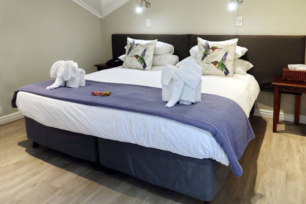 A neatly made bed with white and purple bedding, two pillows with a bird design, and two towels folded on the bed.