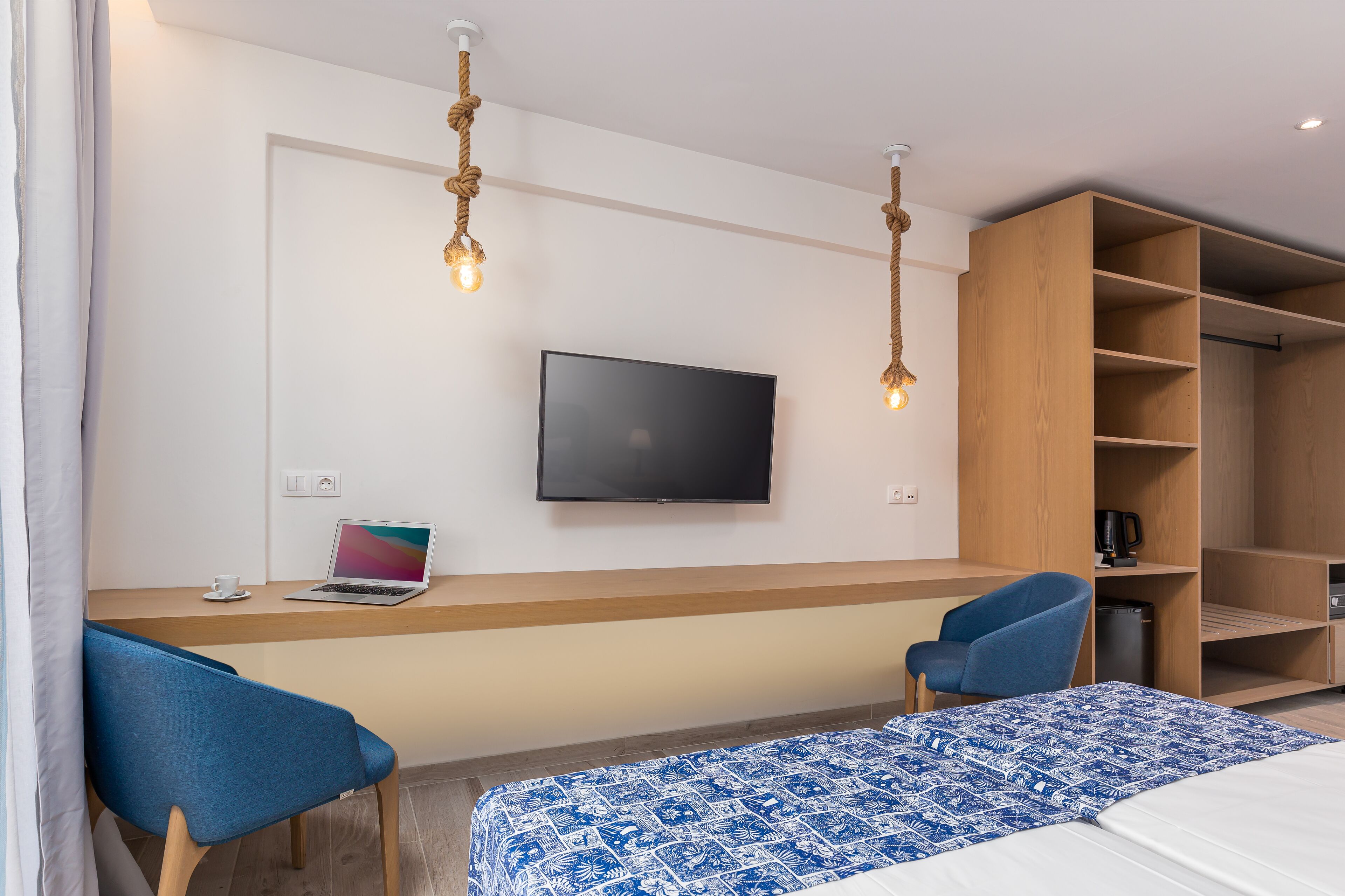 A modern hotel room with a wooden desk, two blue chairs, a bed with a patterned bedspread, a television mounted on the wall, and a wooden wardrobe.