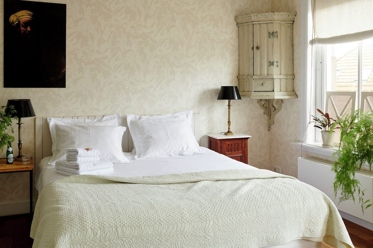 A neatly made bed with white linens and a quilted blanket, a bedside table with a lamp, a small cabinet, and a potted plant by the window.
